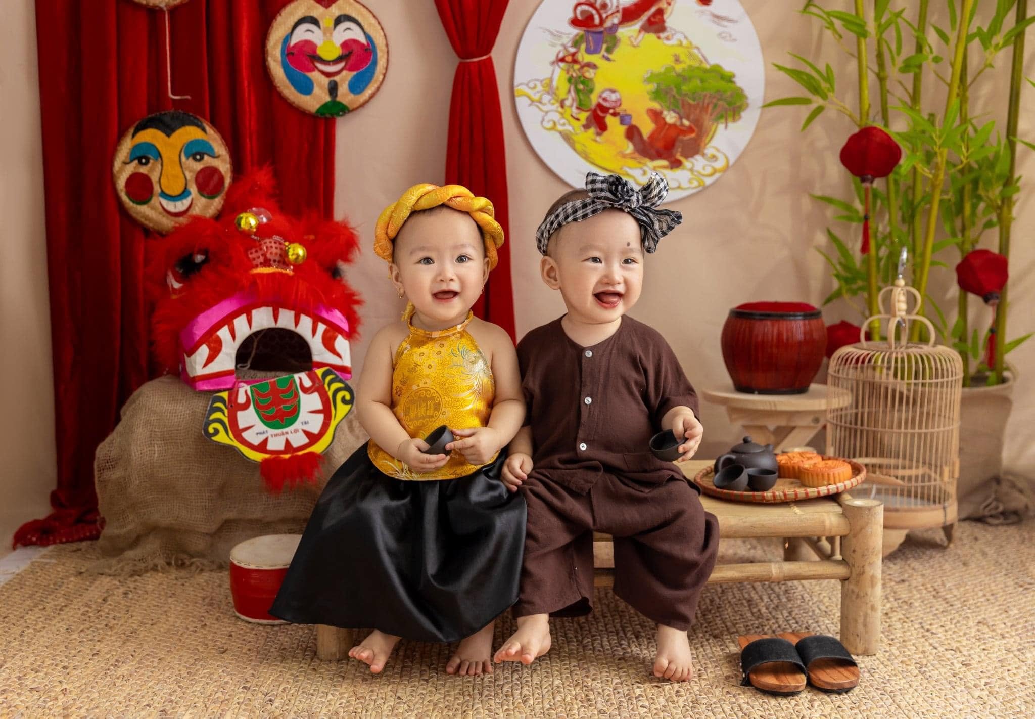 Hai em bé ngồi cạnh nhau bên đầu lân đỏ và mâm bánh nướng đêm Trung thu
