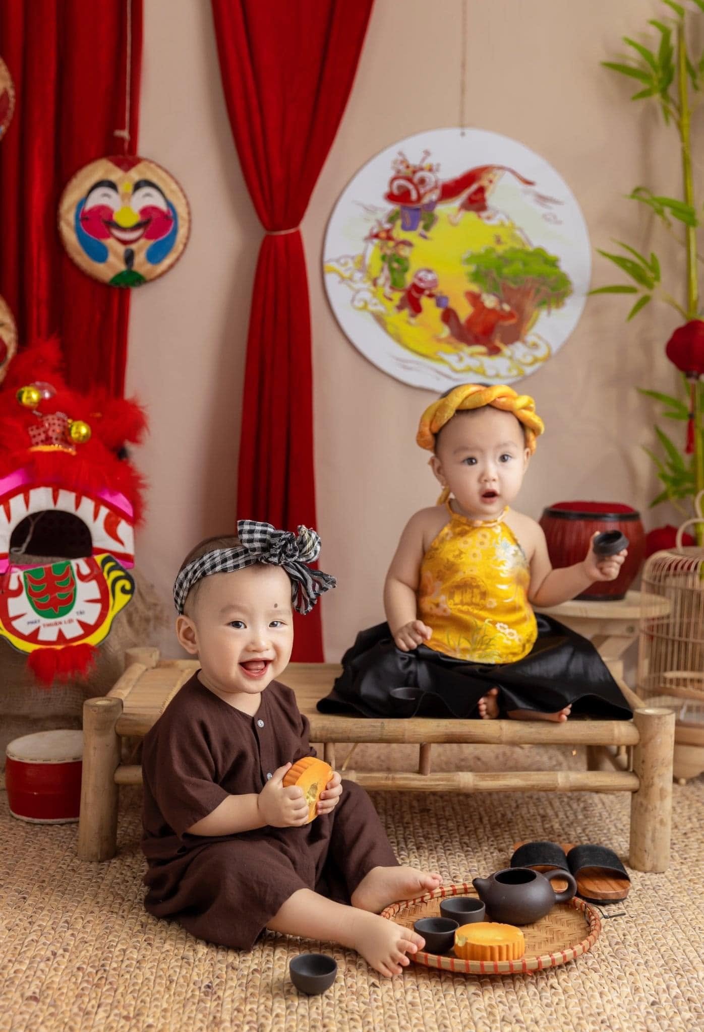 Bé trai áo bà ba nâu cầm chén trà, bé gái yếm vàng ngồi chõng tre bên mâm cỗ Trung thu